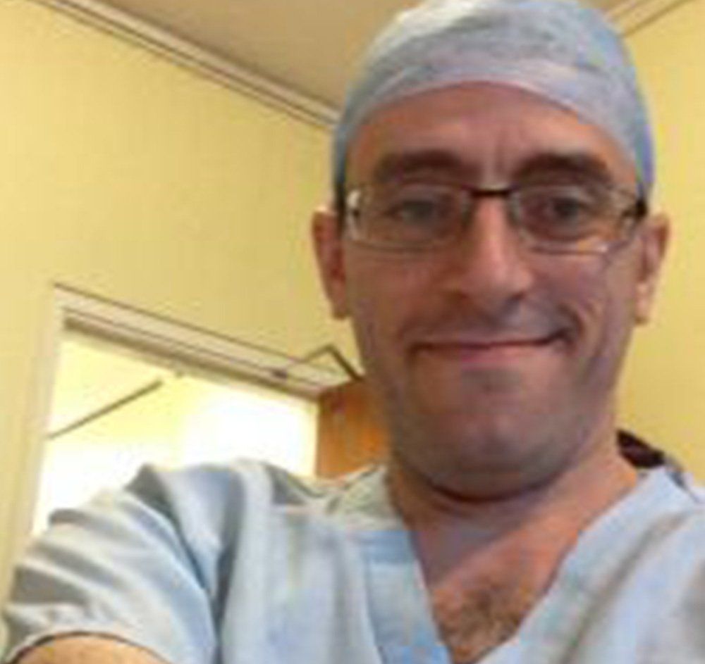 Smiling person in blue medical scrubs and cap, wearing glasses, in a hallway