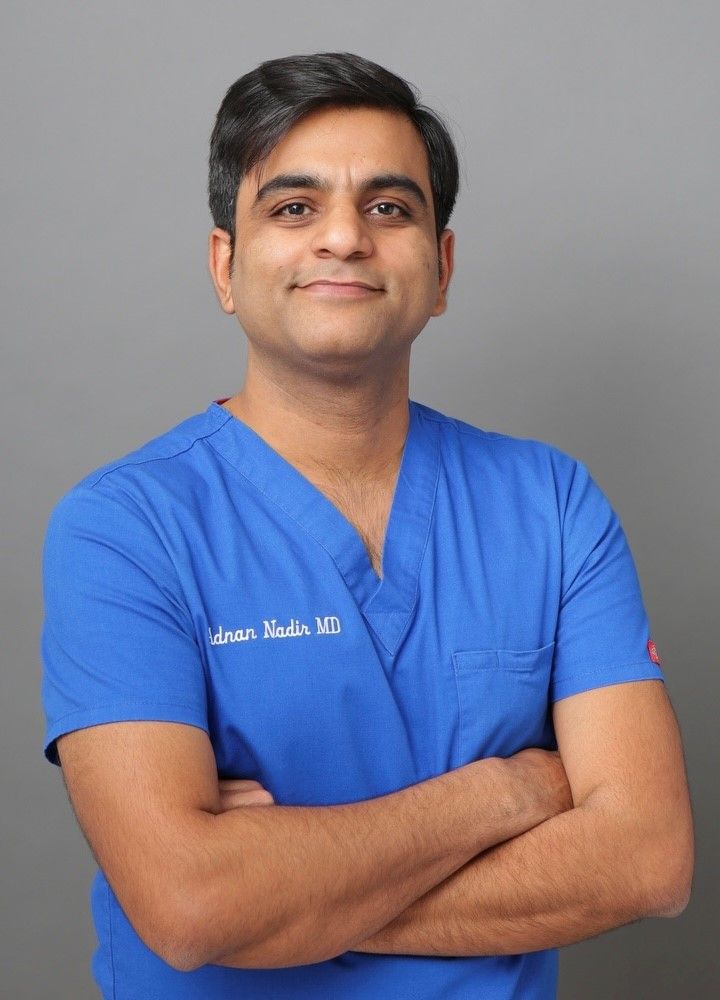 Healthcare professional in blue scrubs standing with arms crossed against a gray background