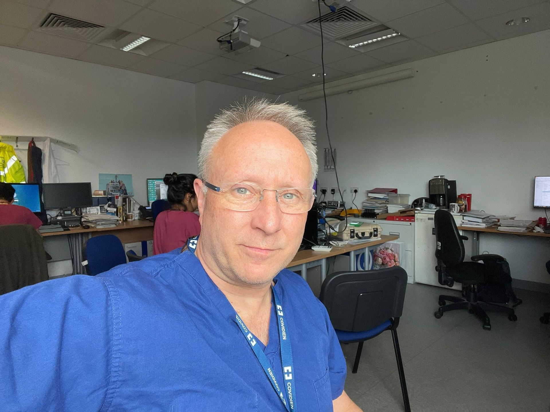 Man in blue shirt taking selfie in a cluttered office with desks, chairs, and computers