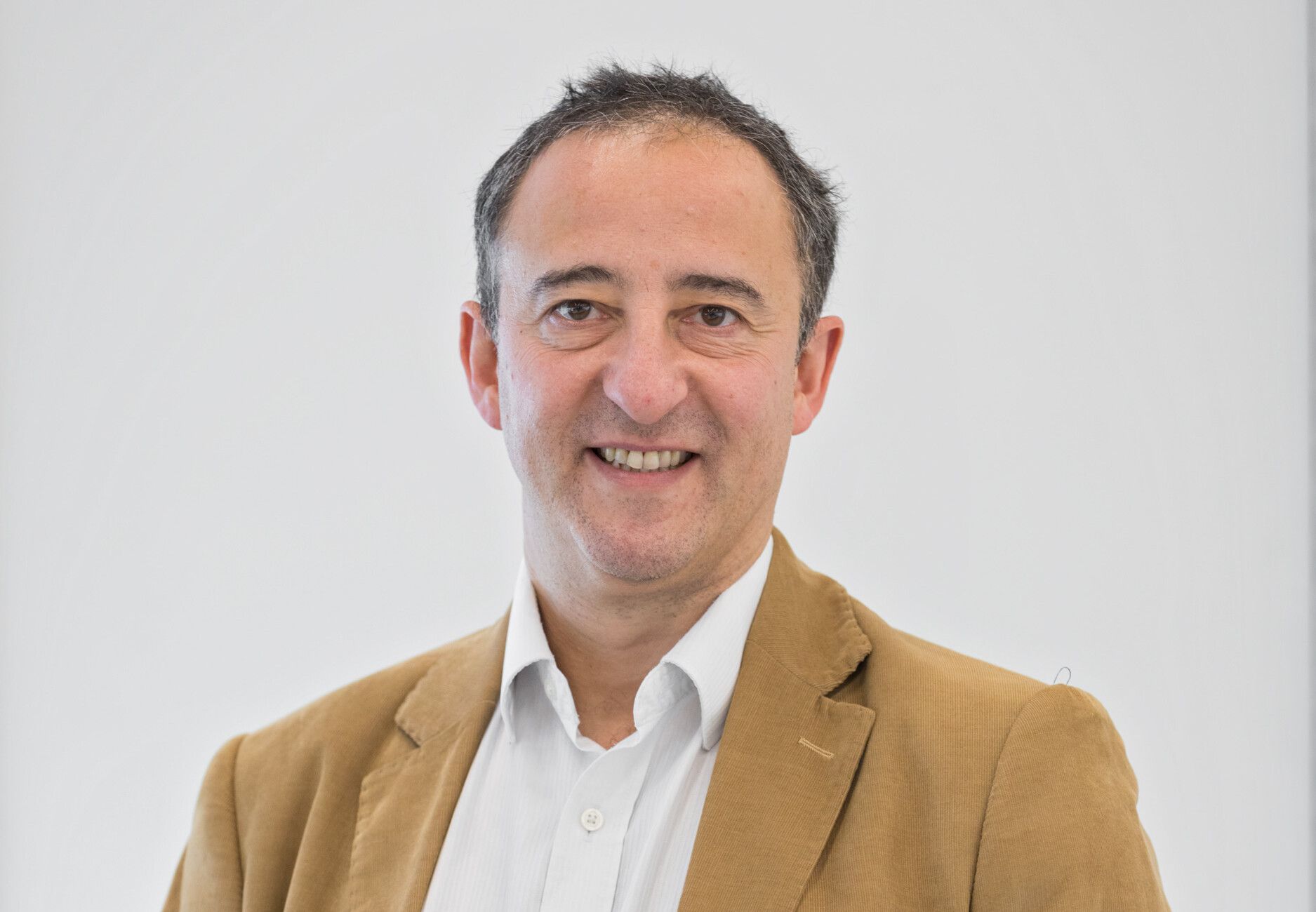Smiling man in a tan blazer and white shirt against a plain light background