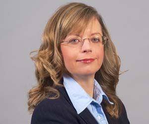 Portrait of a woman wearing glasses, a blue shirt, and a dark blazer against a gray background
