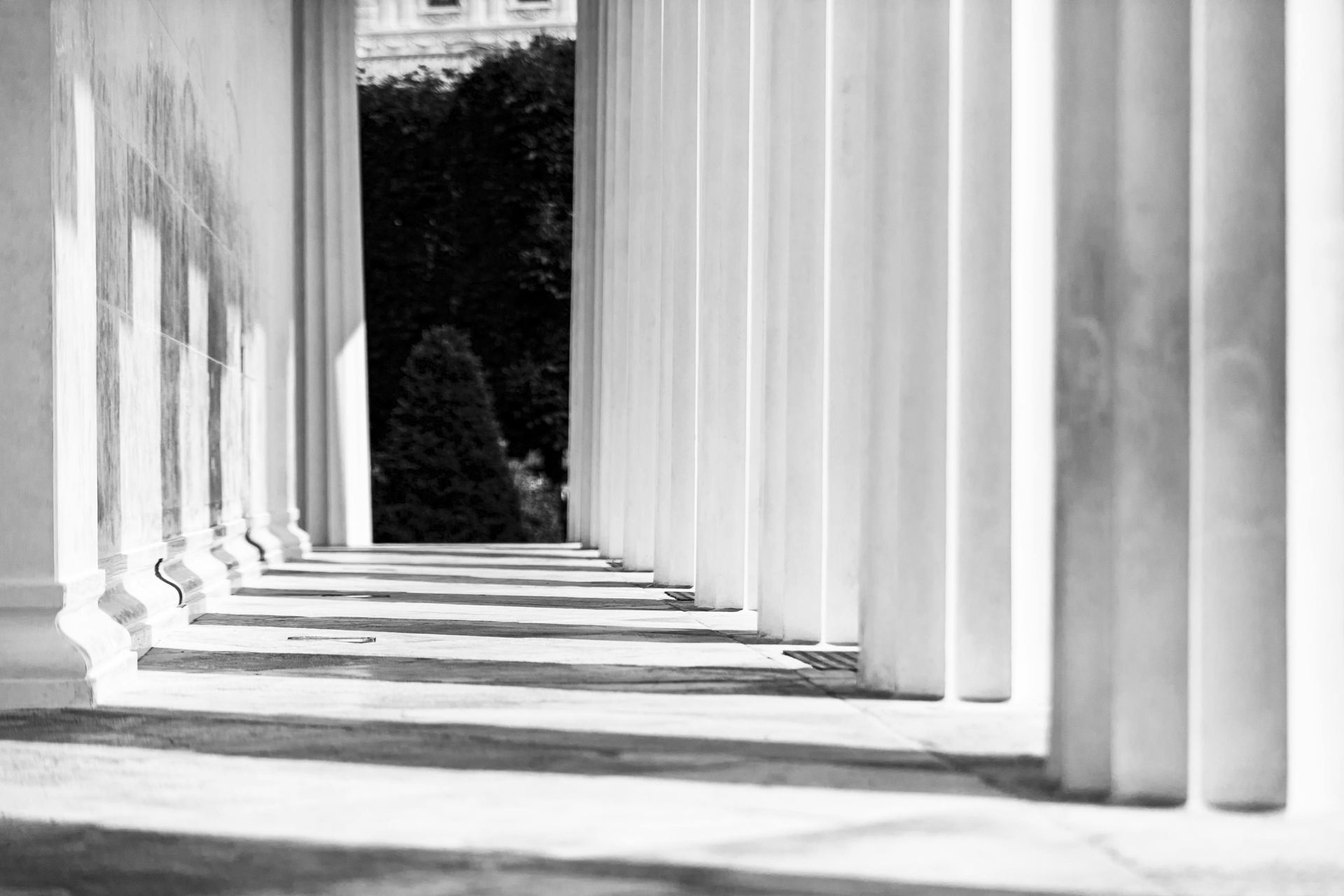 Row of white columns casting long shadows, leading to a dark opening with trees and building visible.