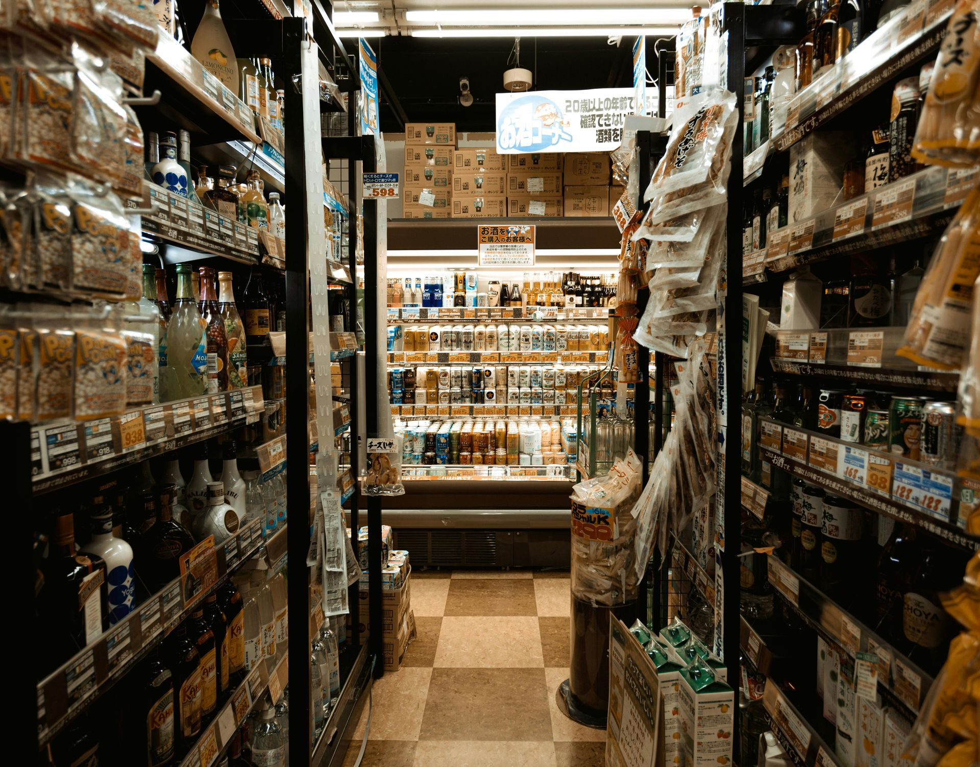Narrow aisle in a store, lined with shelves of products. Checkered floor leads toward a cooler at the end.
