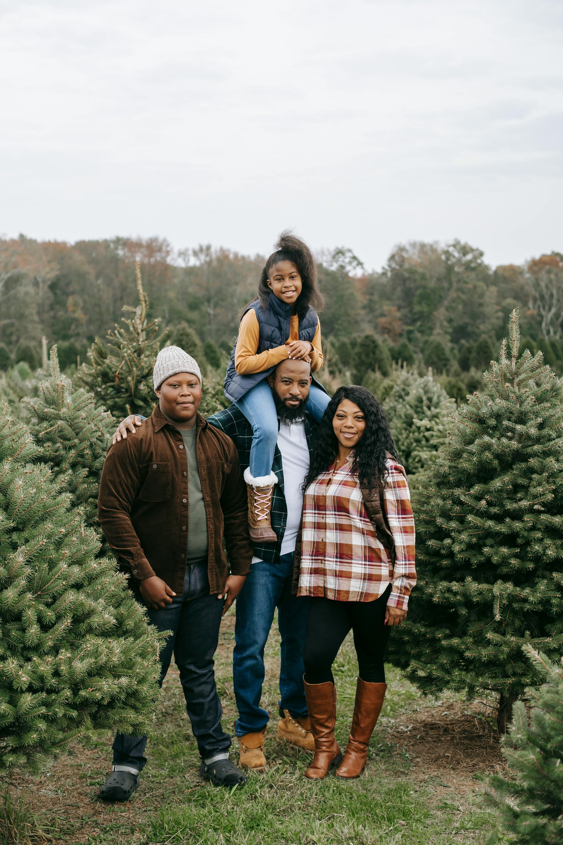 Family poses in a Christmas tree farm; child sits on father's shoulders.