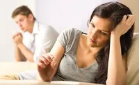 Woman sitting on a couch with a distressed expression while a man sits blurred in the background, both appearing upset