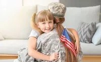 Child hugging a soldier in uniform while holding a small American flag on a couch