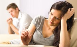 Woman in gray shirt sits upset on a couch while a blurred man sits in the background.