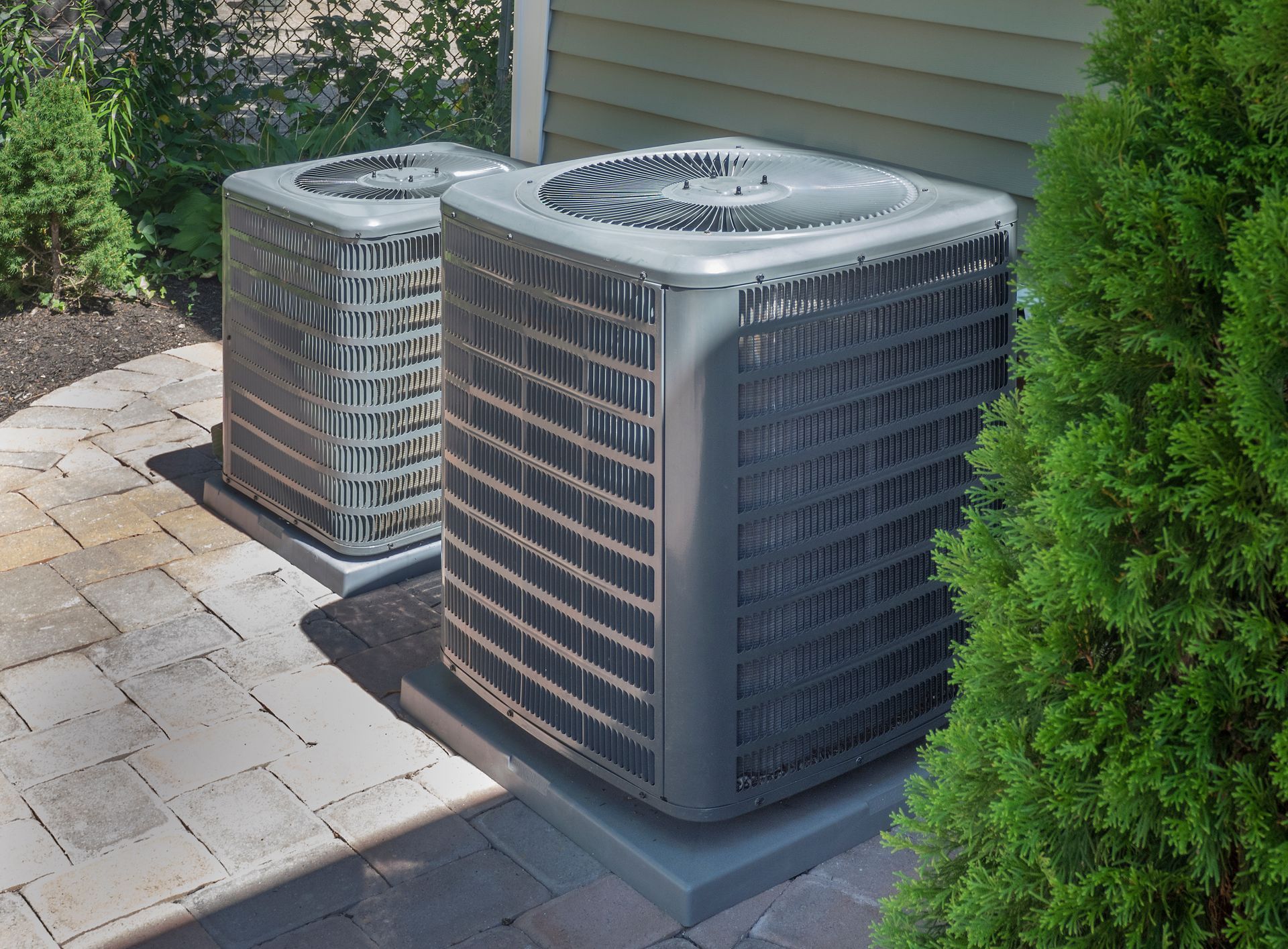 Two outdoor air conditioning units beside a house, near a paved walkway and shrubs.