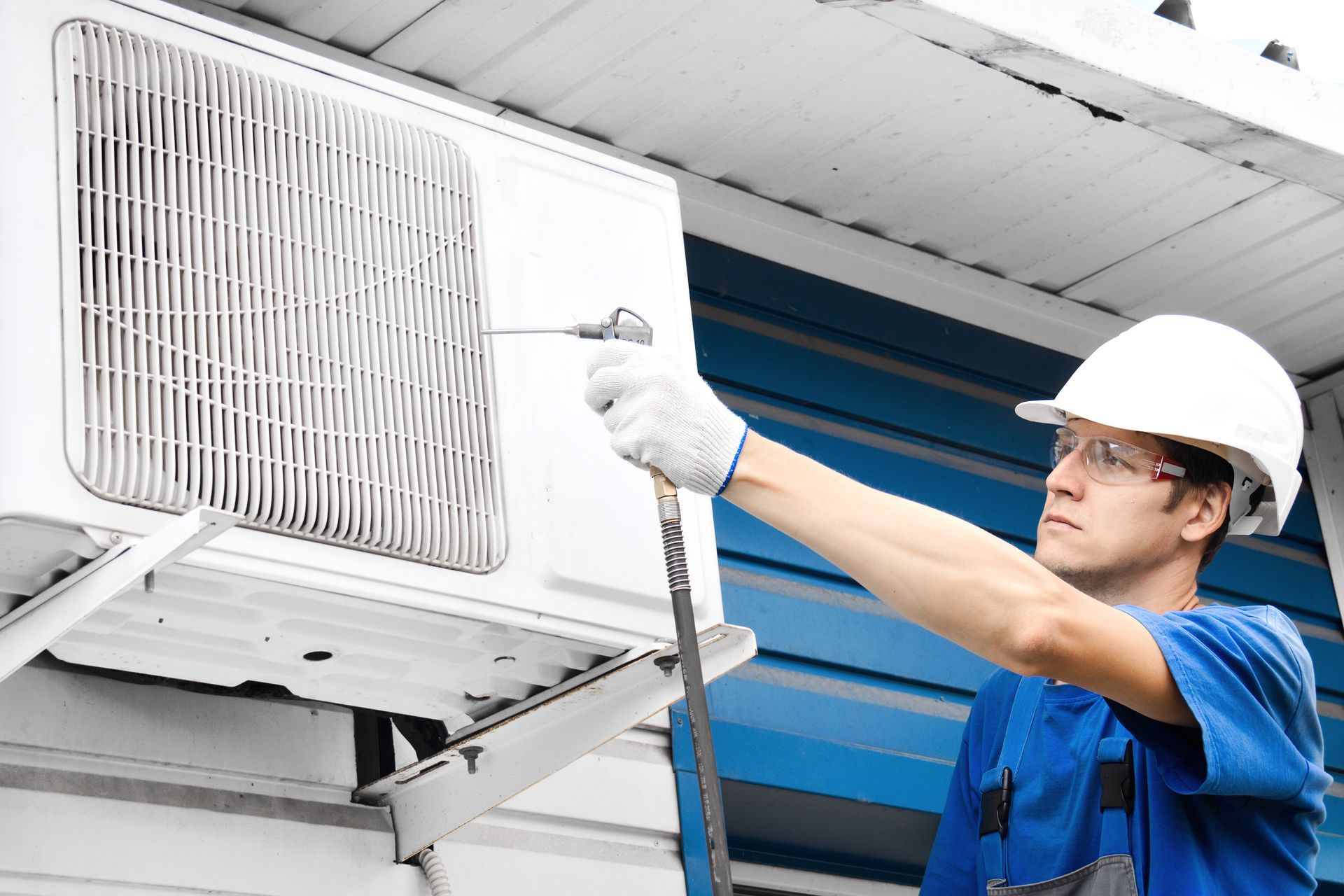 Technician in blue uniform and hard hat servicing an outdoor air conditioner unit on a building wall