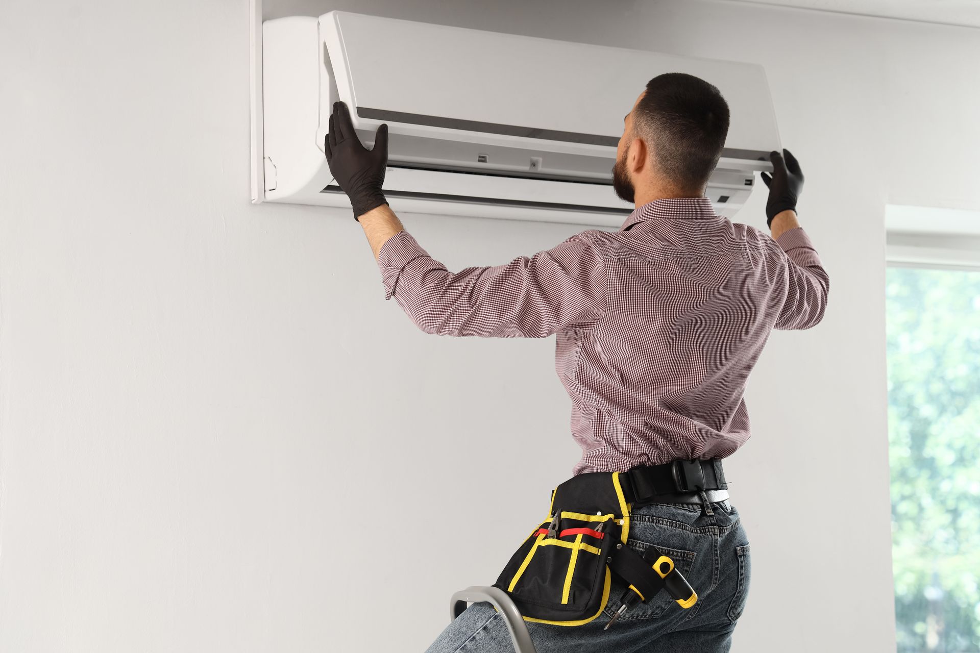 Technician installing a wall-mounted air conditioner indoors, using a tool belt and gloves