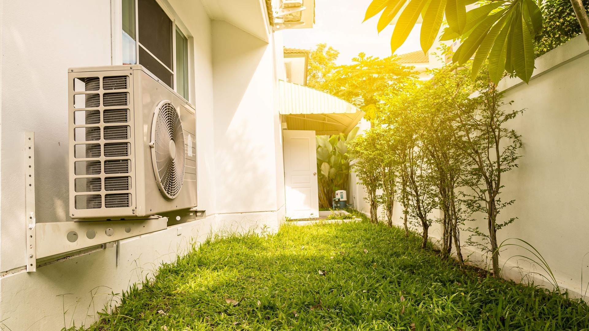 Outdoor air conditioning unit beside a grassy walkway and white wall, with bright sunlight and trees nearby