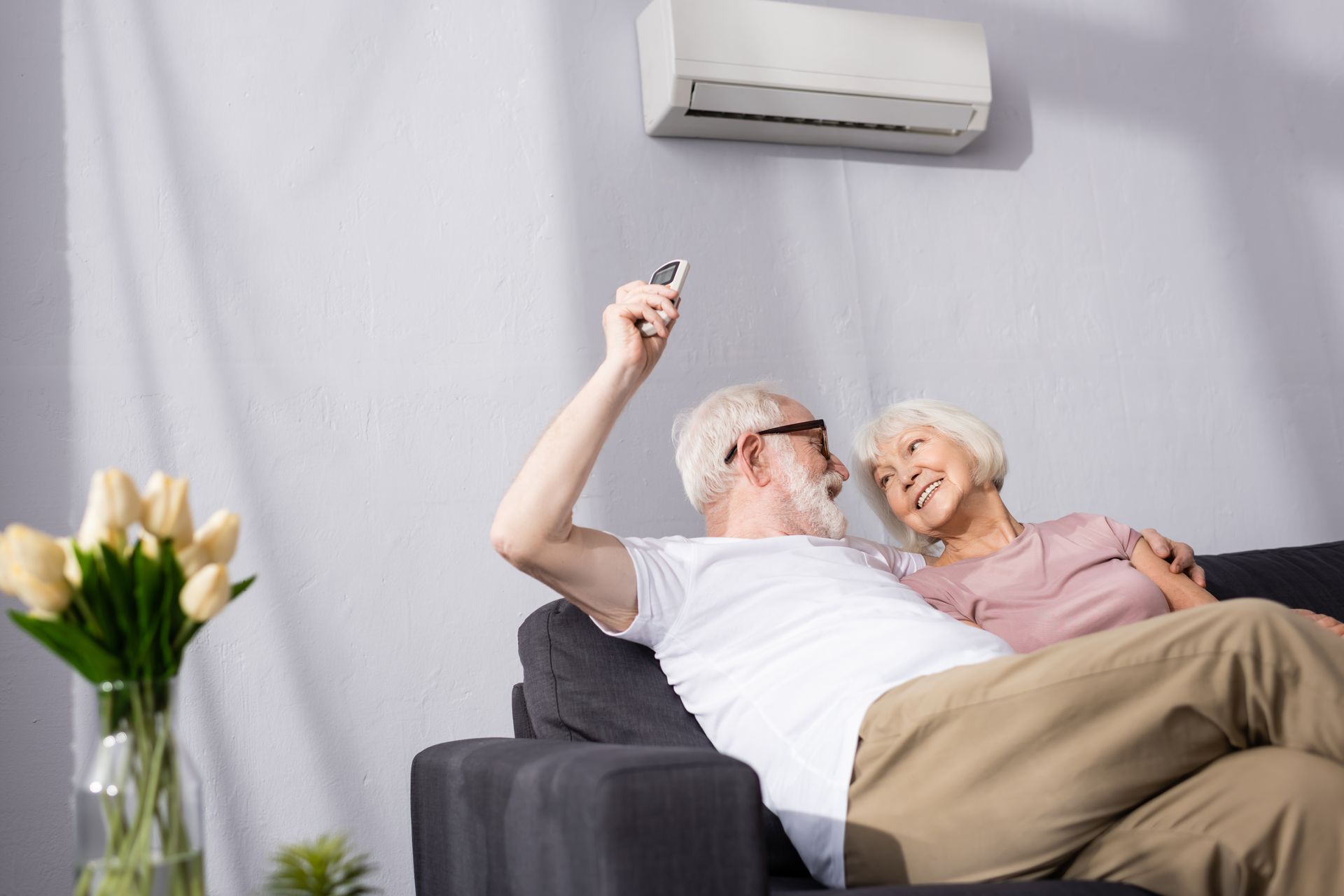 Older couple smiling on a sofa, taking a selfie in a bright living room with a wall-mounted air conditioner.