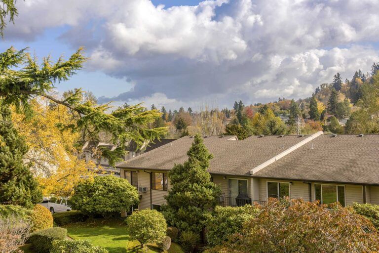 Suburban house with a brown roof, green shrubs, and autumn trees under a cloudy sky
