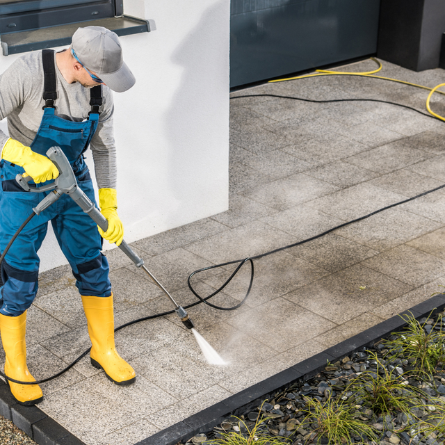 Person in overalls and rubber boots pressure washing a concrete patio.