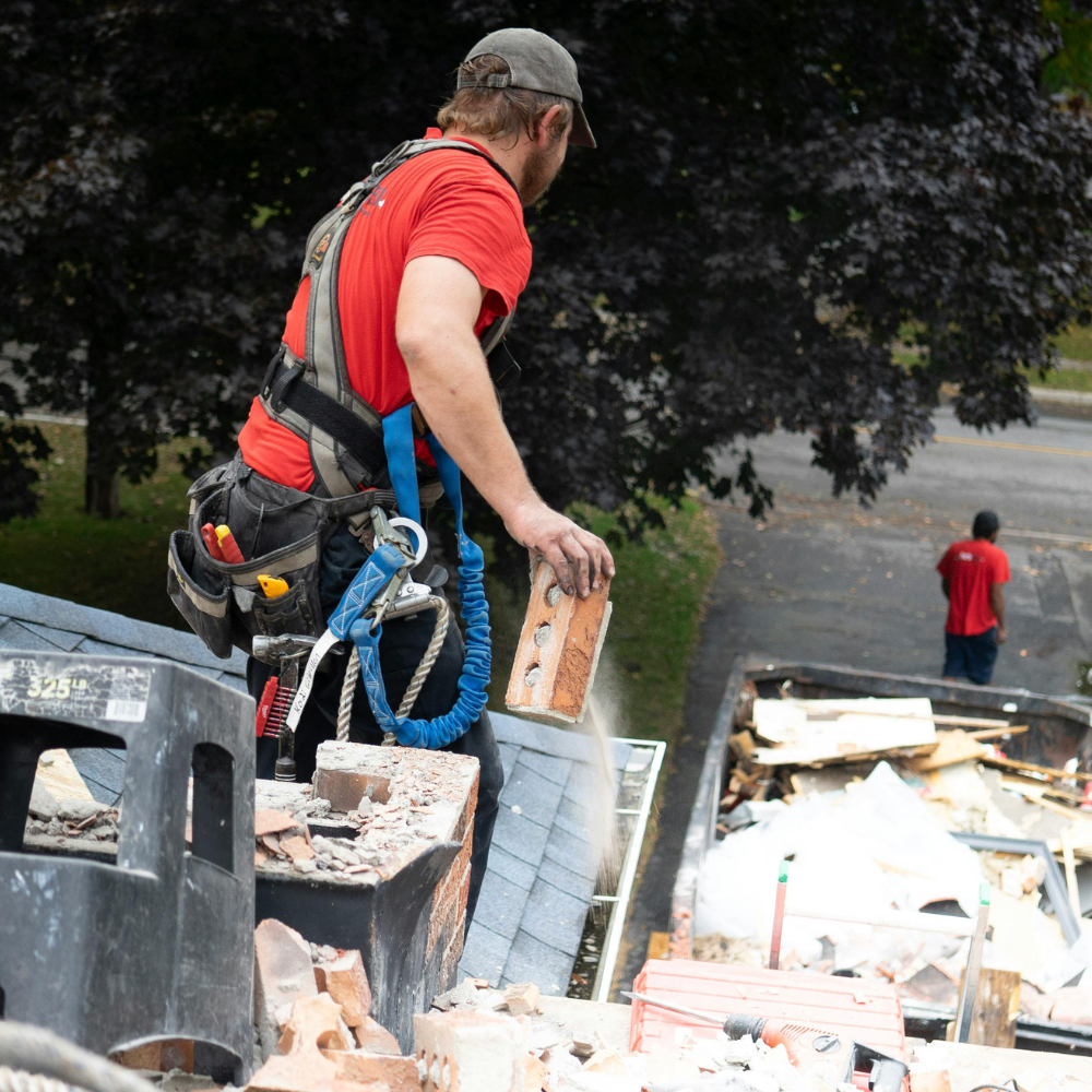 Roofer on a roof, in safety harness, holding brick; another worker in the background near debris and a dumpster.