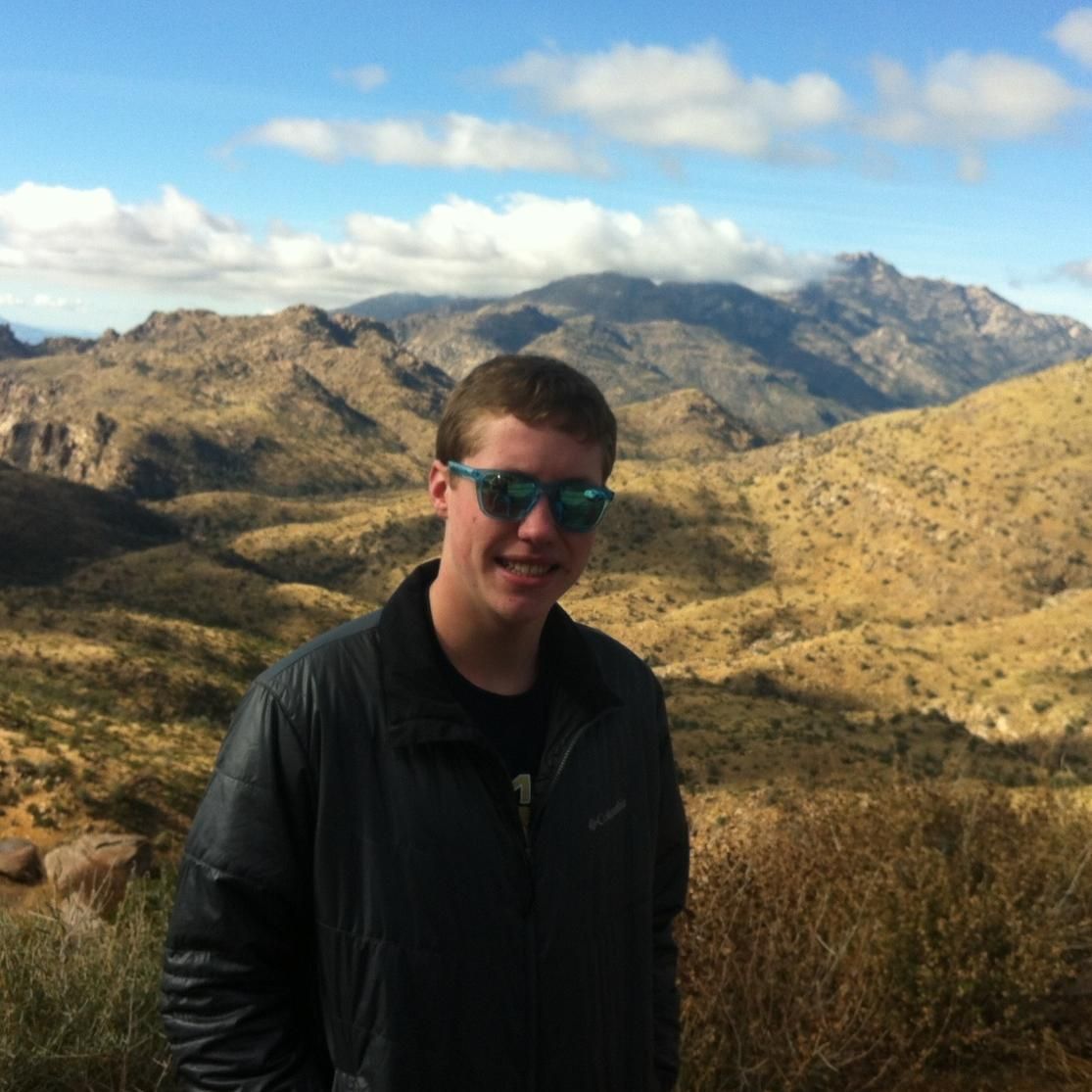 Person in sunglasses and black jacket stands in a dry mountain landscape under a cloudy sky.