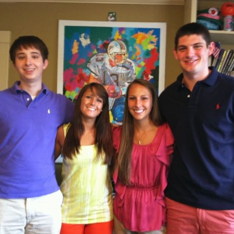 Four smiling students posing indoors in front of a colorful painting.