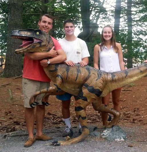 Three people posing with a large dinosaur model in a wooded outdoor setting.