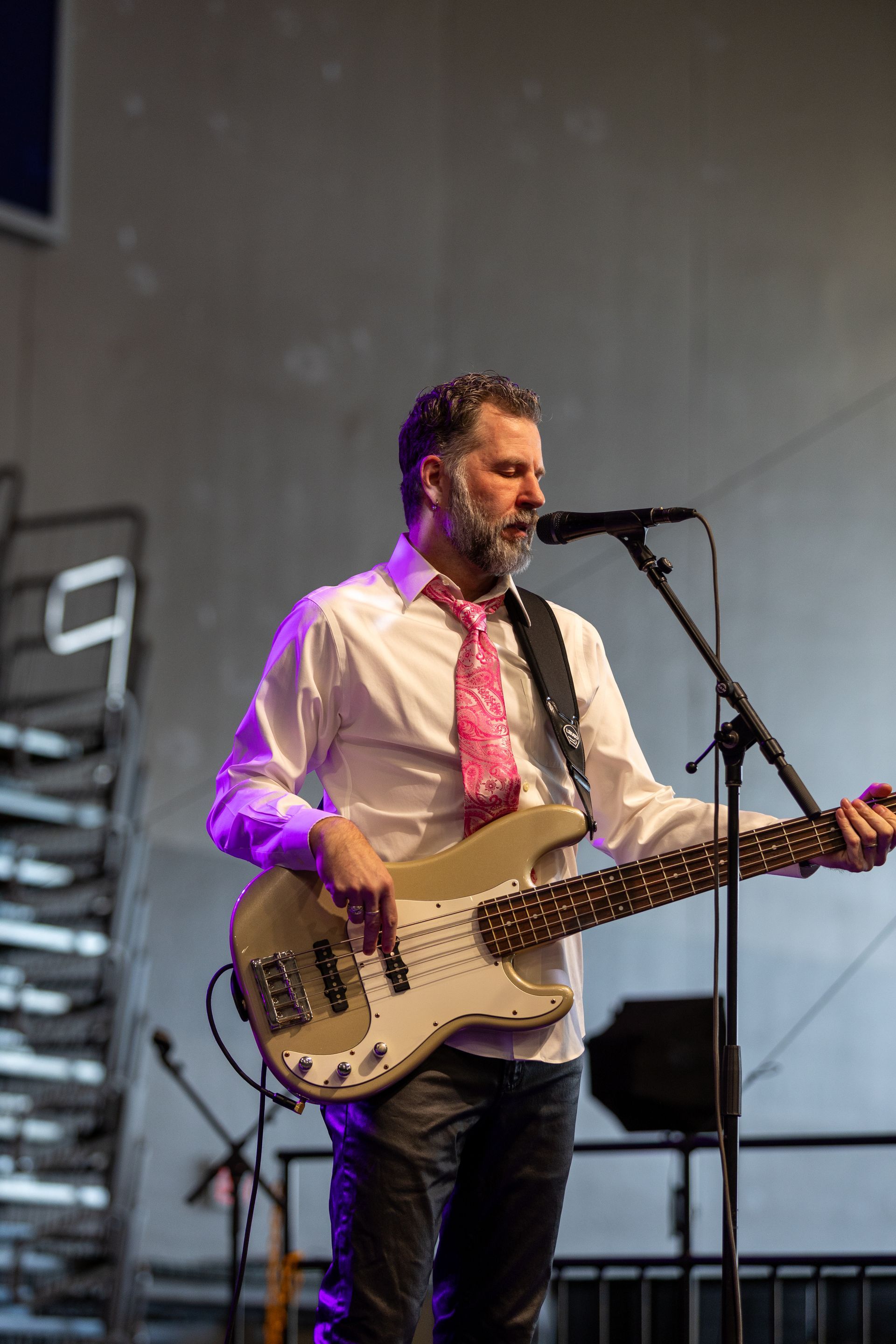 Musician singing into a microphone while playing a white bass guitar onstage, wearing a white shirt and pink tie