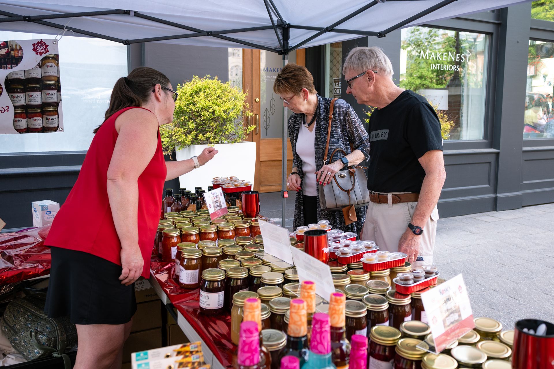 People browsing jars of preserves at a market stall under a canopy.