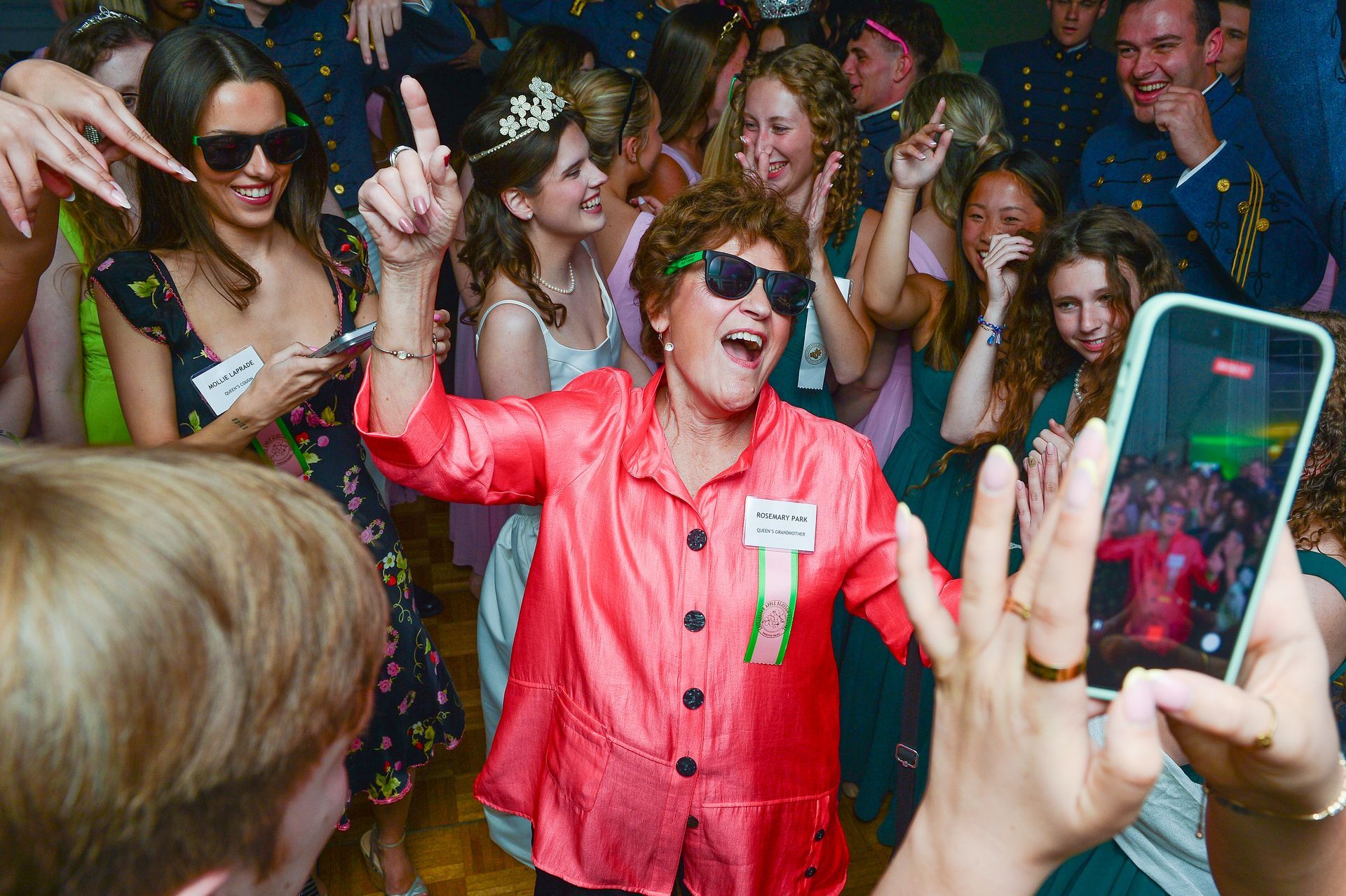 Crowd cheering at an event as a person in a bright pink jacket raises a finger and smiles while others film with phones
