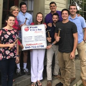 Group of smiling people holding a Full Circle sign and tools on a porch, standing together for a team photo