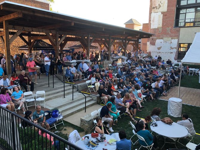 Crowd seated outdoors at a public event, with a stage under a wooden pavilion and tables in the foreground