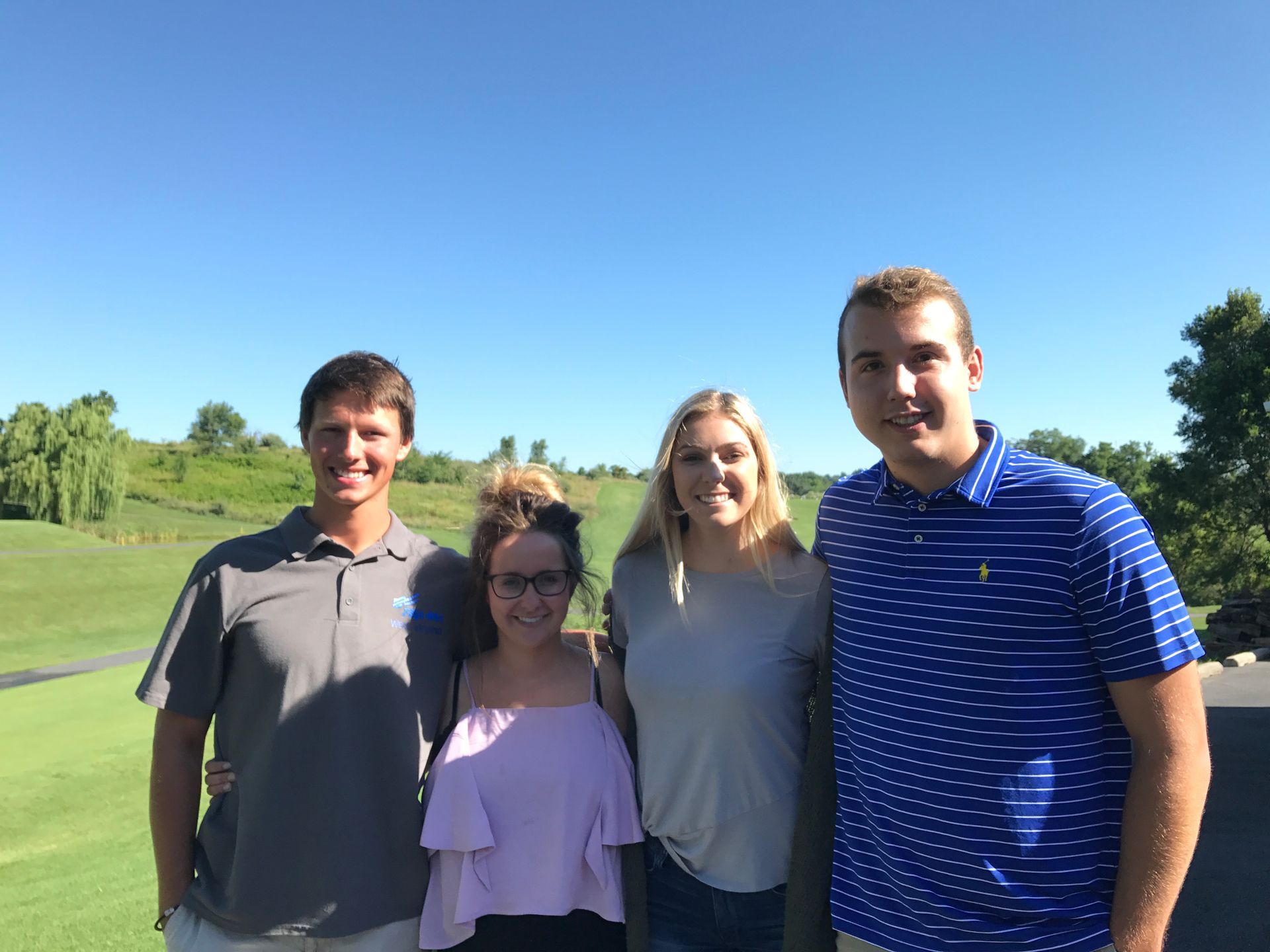 Four people standing outdoors on a sunny grassy field under a blue sky.