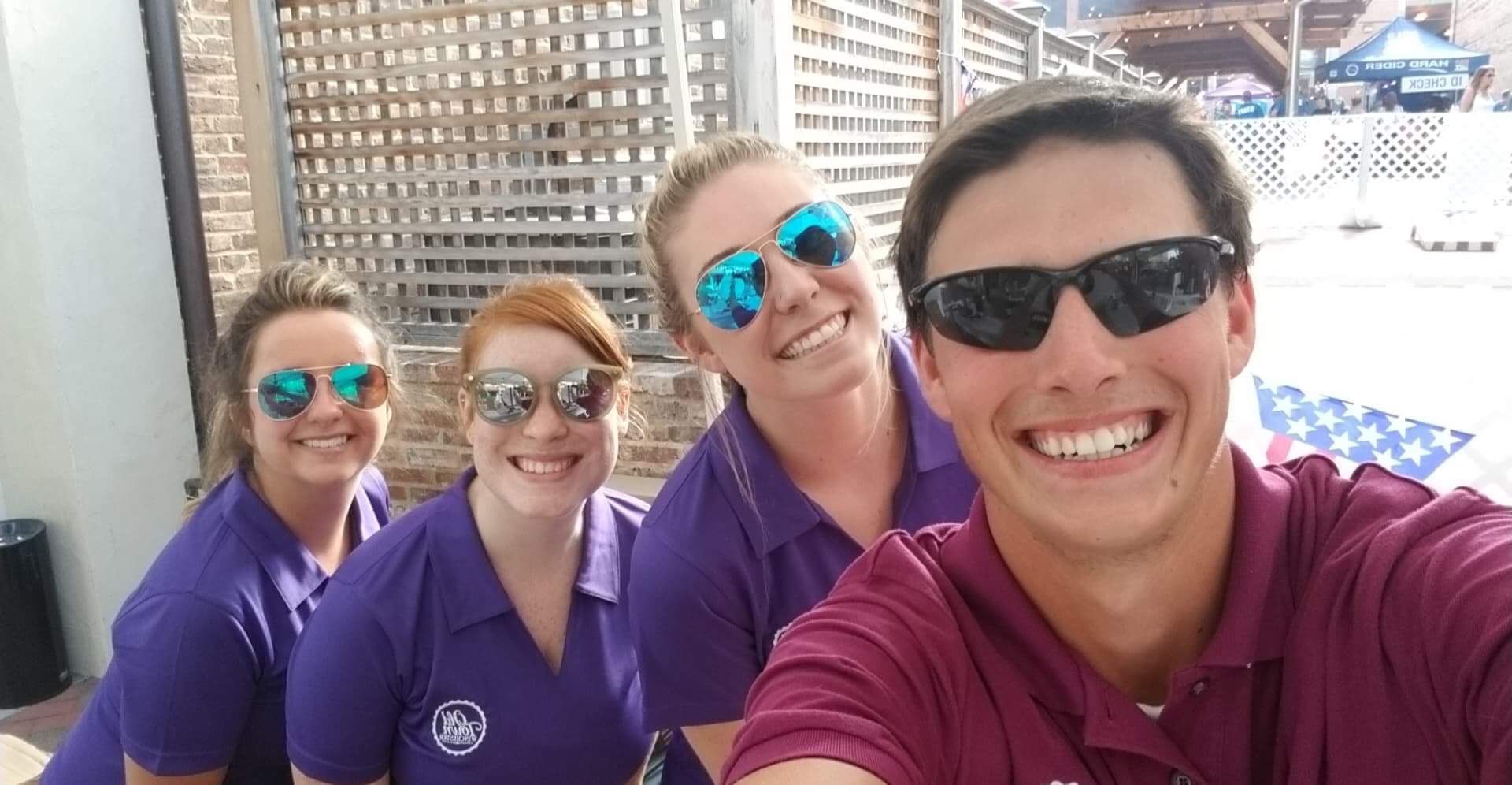 Four smiling coworkers in purple shirts pose for a selfie outdoors in bright sunlight.