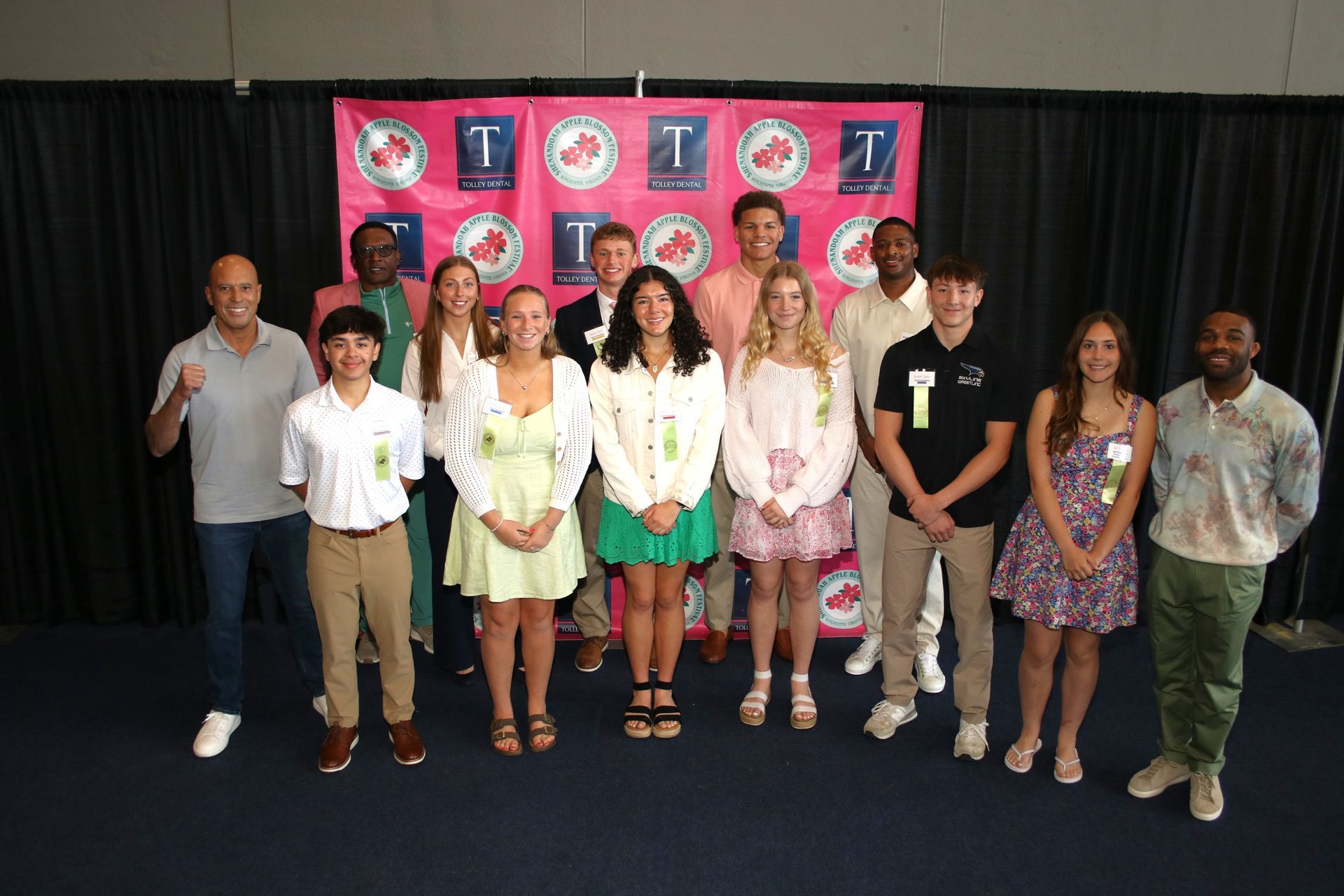 Group of people posing on stage in front of a pink backdrop, holding microphones and wearing event badges