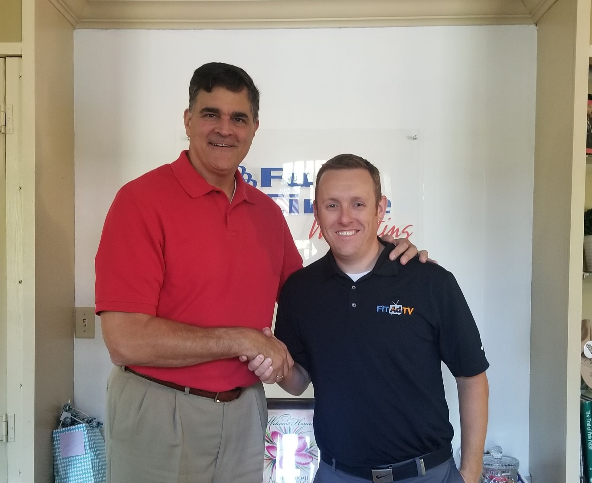 Two men standing indoors, shaking hands and smiling, one in a red shirt and one in a navy polo.