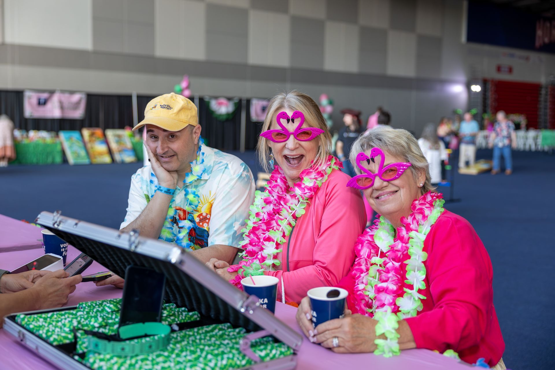 Three smiling people in pink and floral leis at a table, wearing heart-shaped glasses and hats indoors Jimmy Buffett