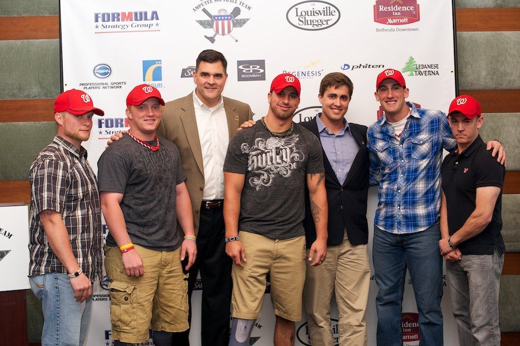 Seven men posing in front of a sponsor backdrop, several wearing red Nationals caps and casual shirts, at an event.