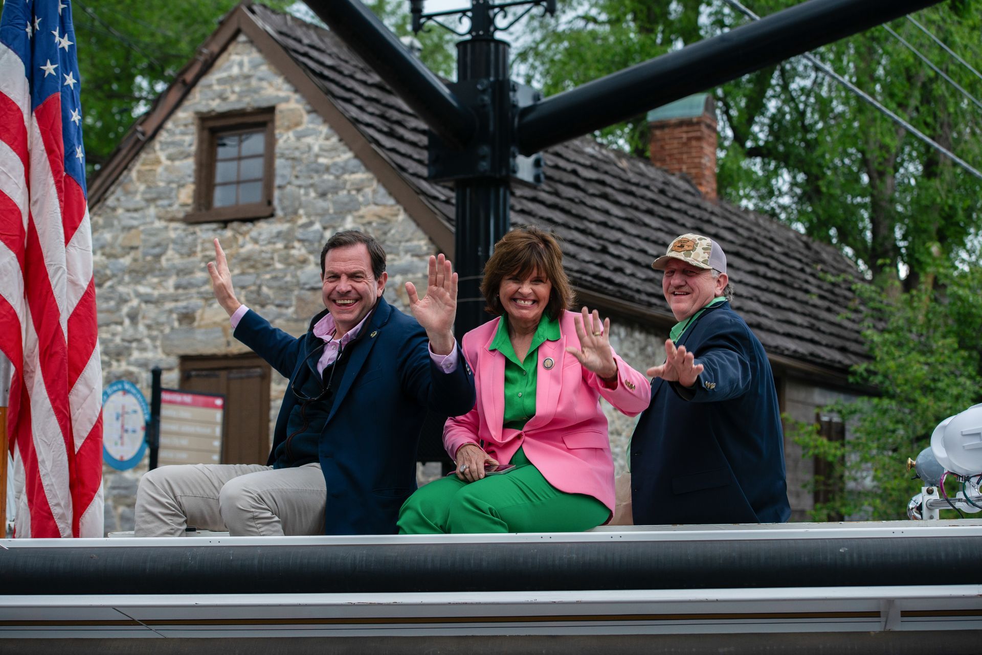 Three people wave from an open vehicle in front of a chalet-style house and trees.
