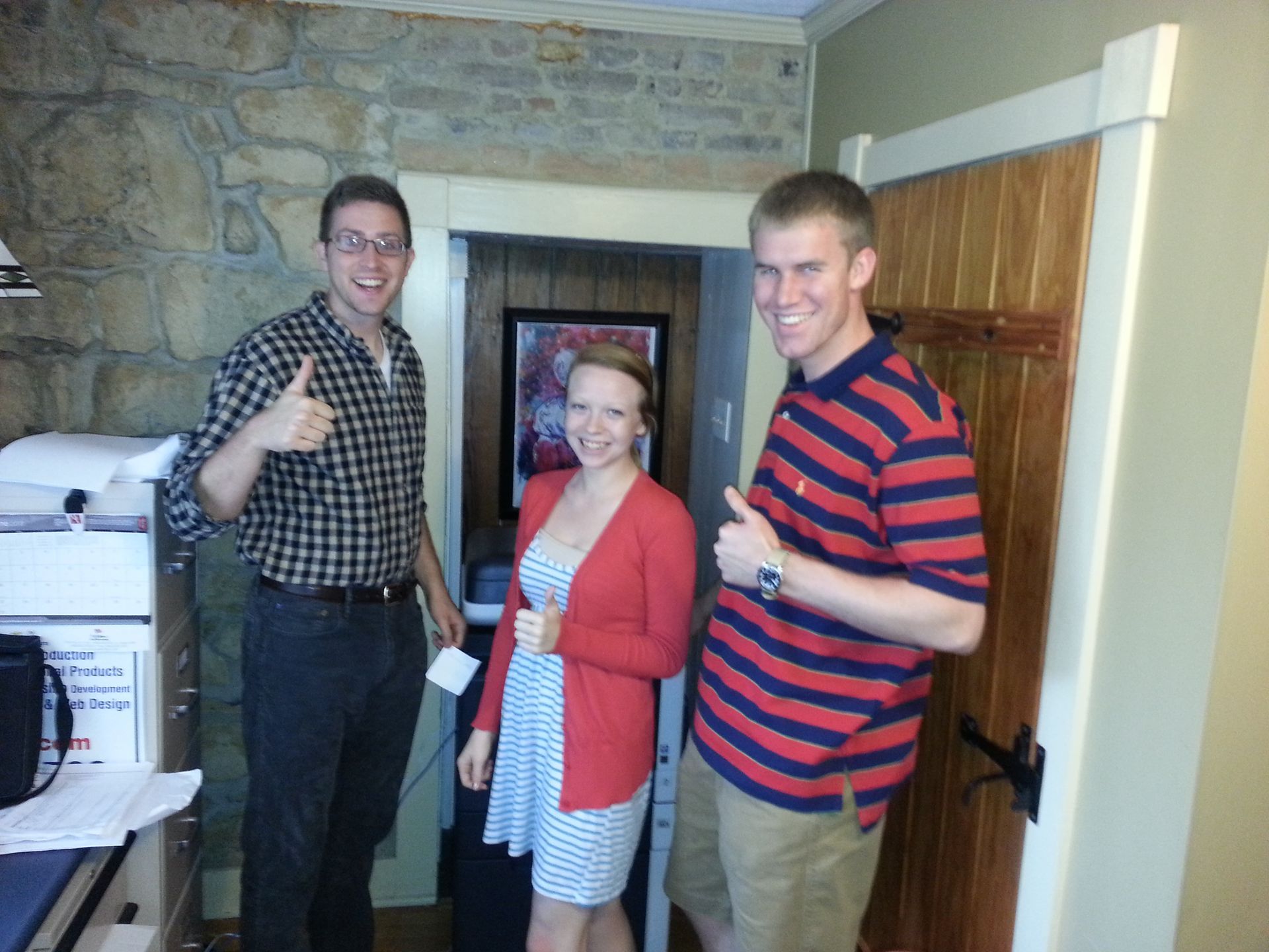 Three people smiling and posing indoors near a doorway, two giving thumbs-up.