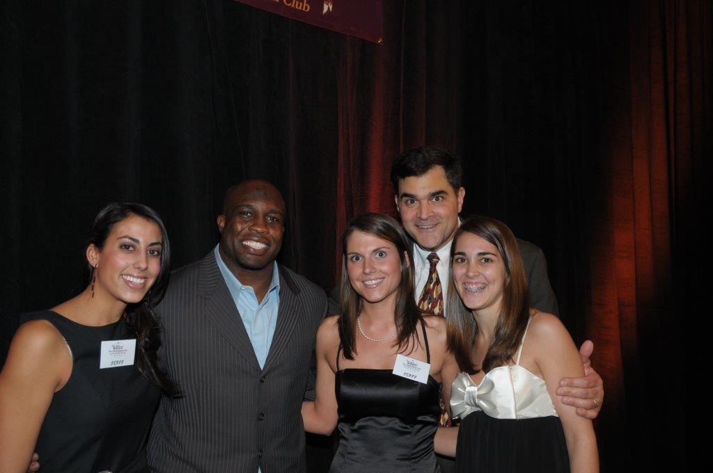 Five people posing together at a dimly lit event, smiling in formal attire.