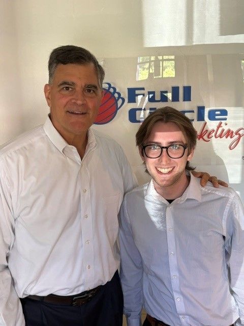 Two men smiling in white dress shirts standing in front of a Full Circle Marketing sign