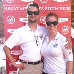 Two people in white polo shirts posing in front of a red event backdrop, both wearing sunglasses. Intern