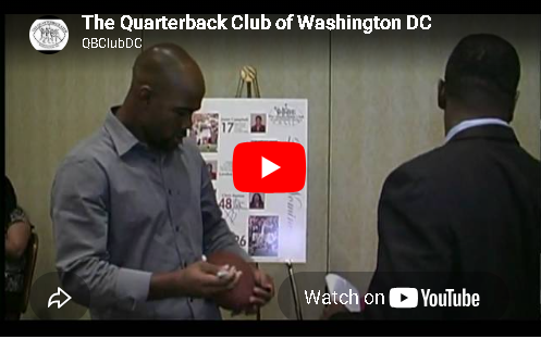Two men stand beside a wall poster with a YouTube play button overlay in a conference room