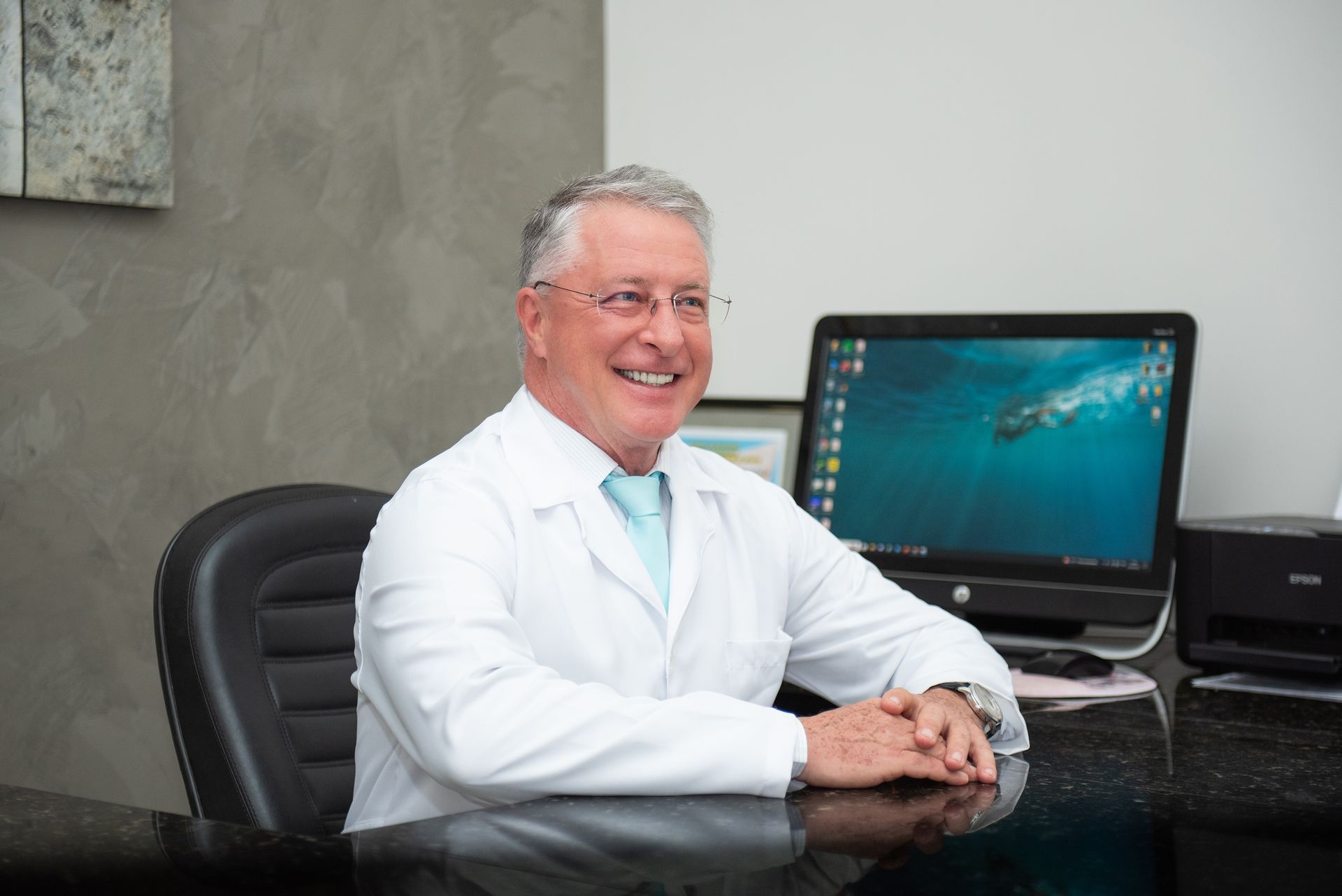 Médico sorridente de jaleco branco sentado à mesa com computador em um consultório.