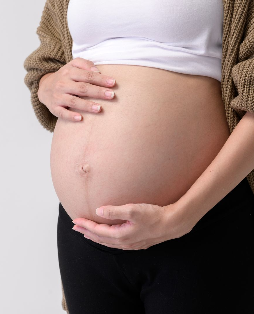 Mulher grávida segurando a barriga nua com as duas mãos, vestindo uma blusa branca e um cardigã marrom.