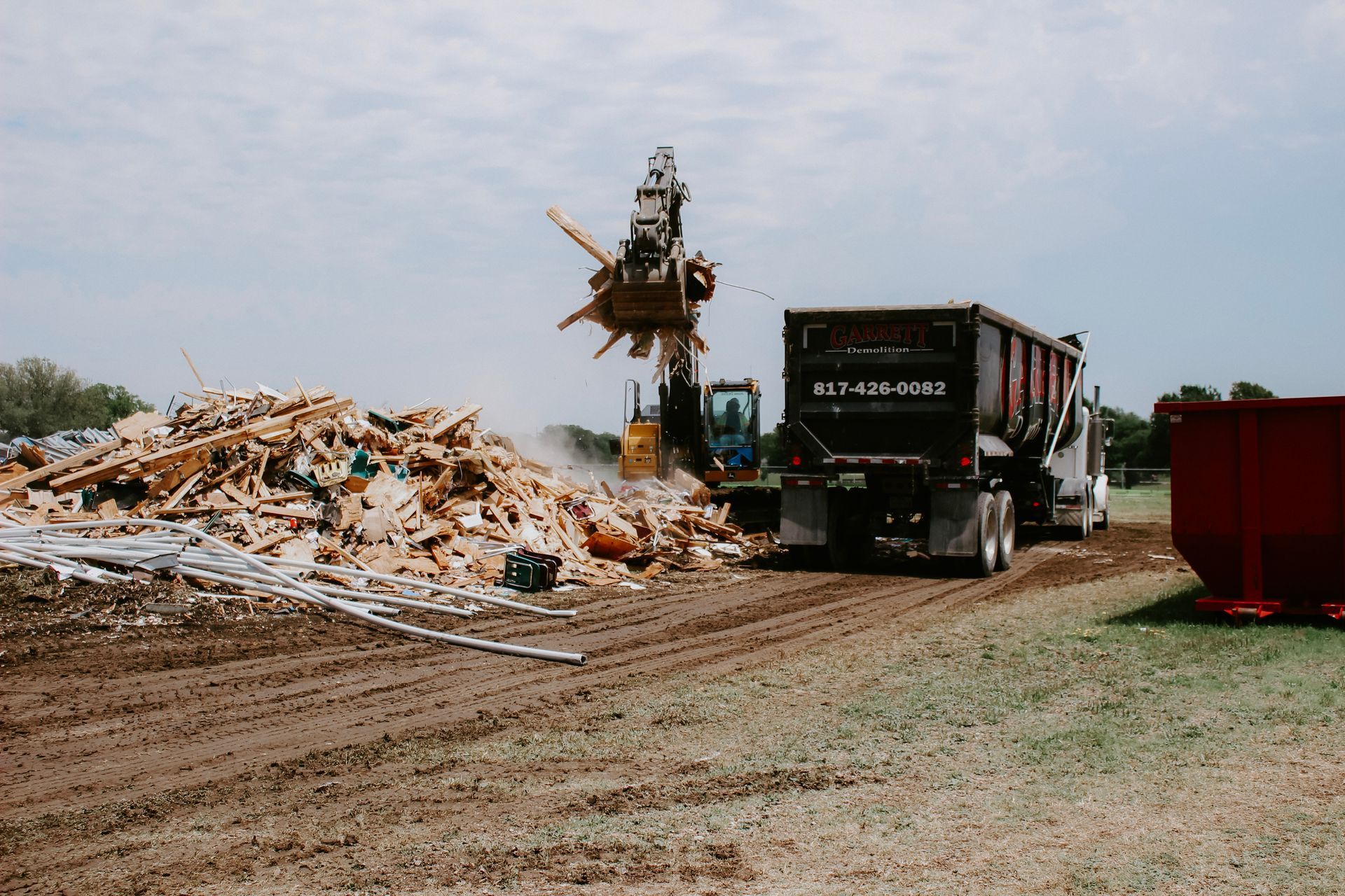 A dump truck is driving down a dirt road next to a pile of wood.
