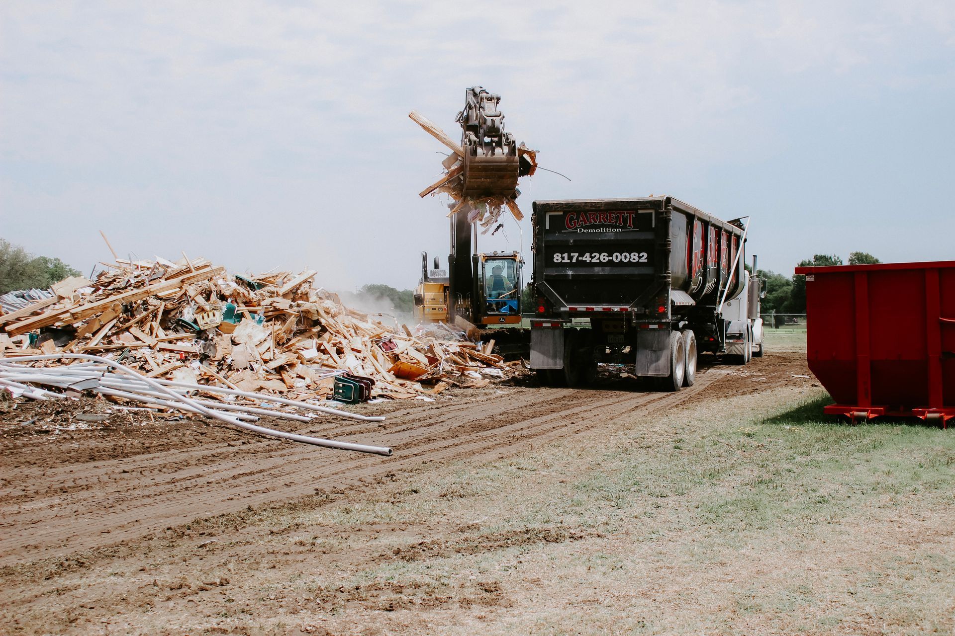 A dump truck is driving down a dirt road next to a pile of wood.