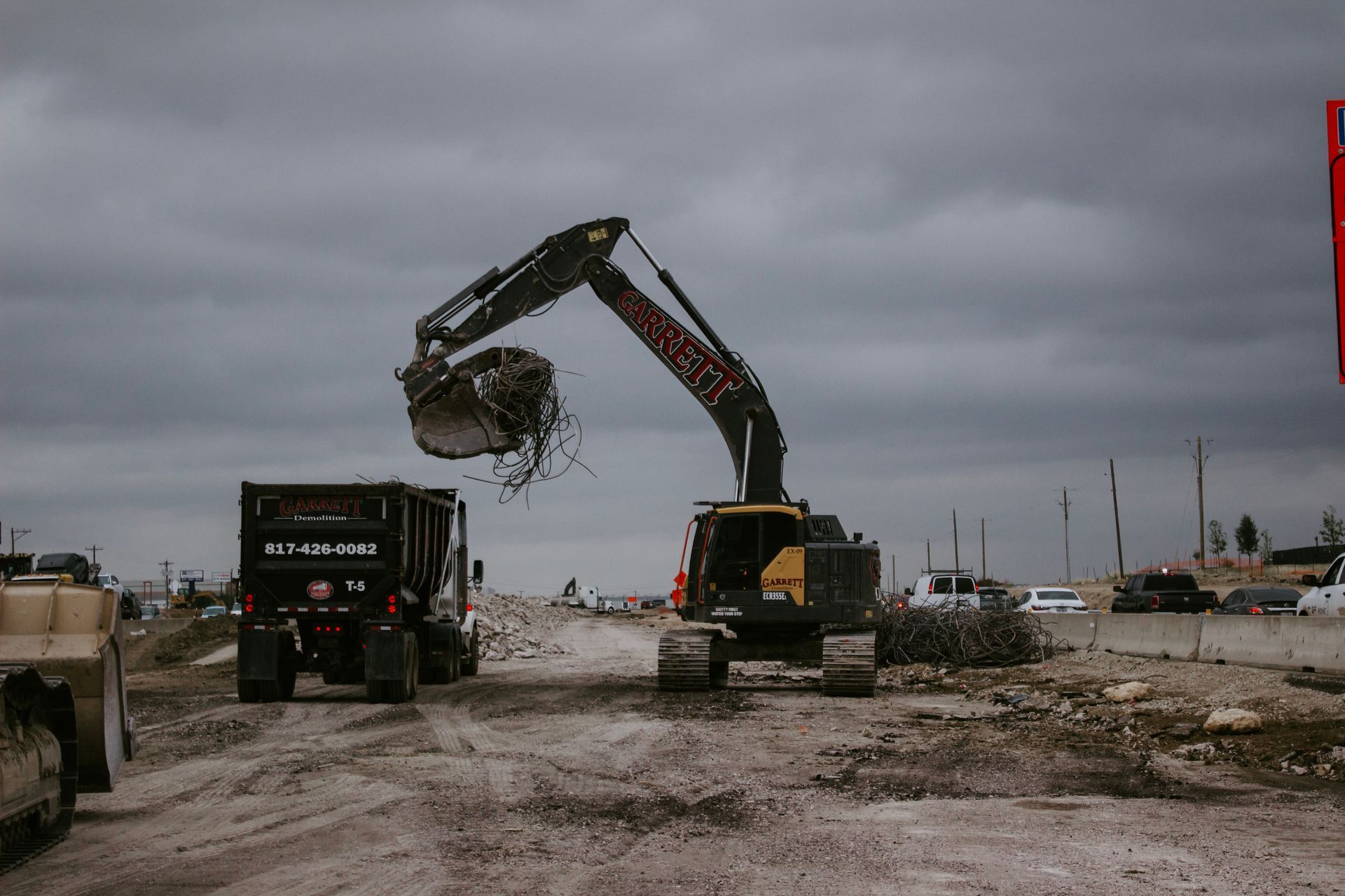 A large excavator is loading a dumpster in a dirt lot.