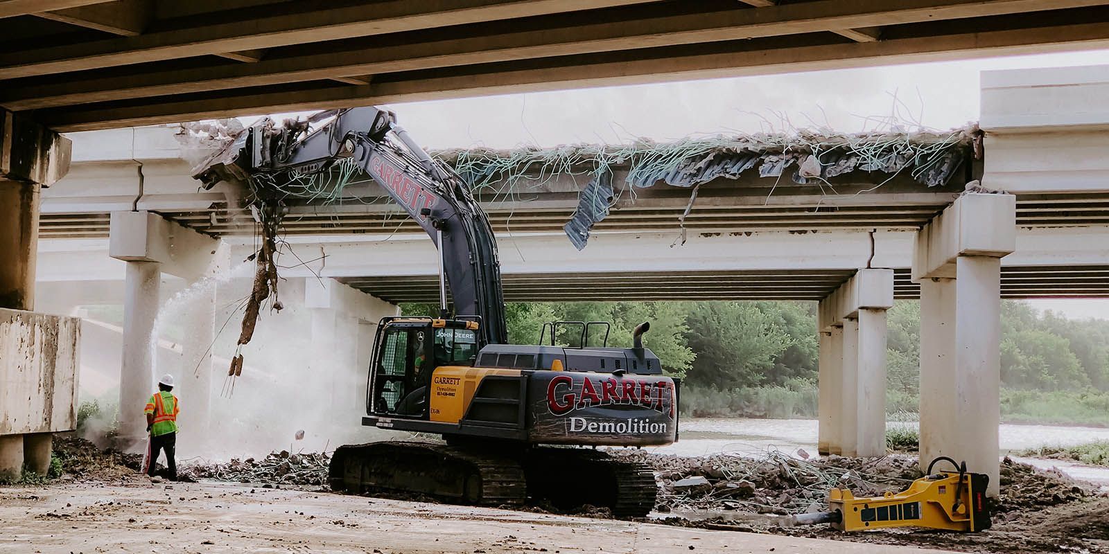 A large excavator is demolishing a bridge under a highway.