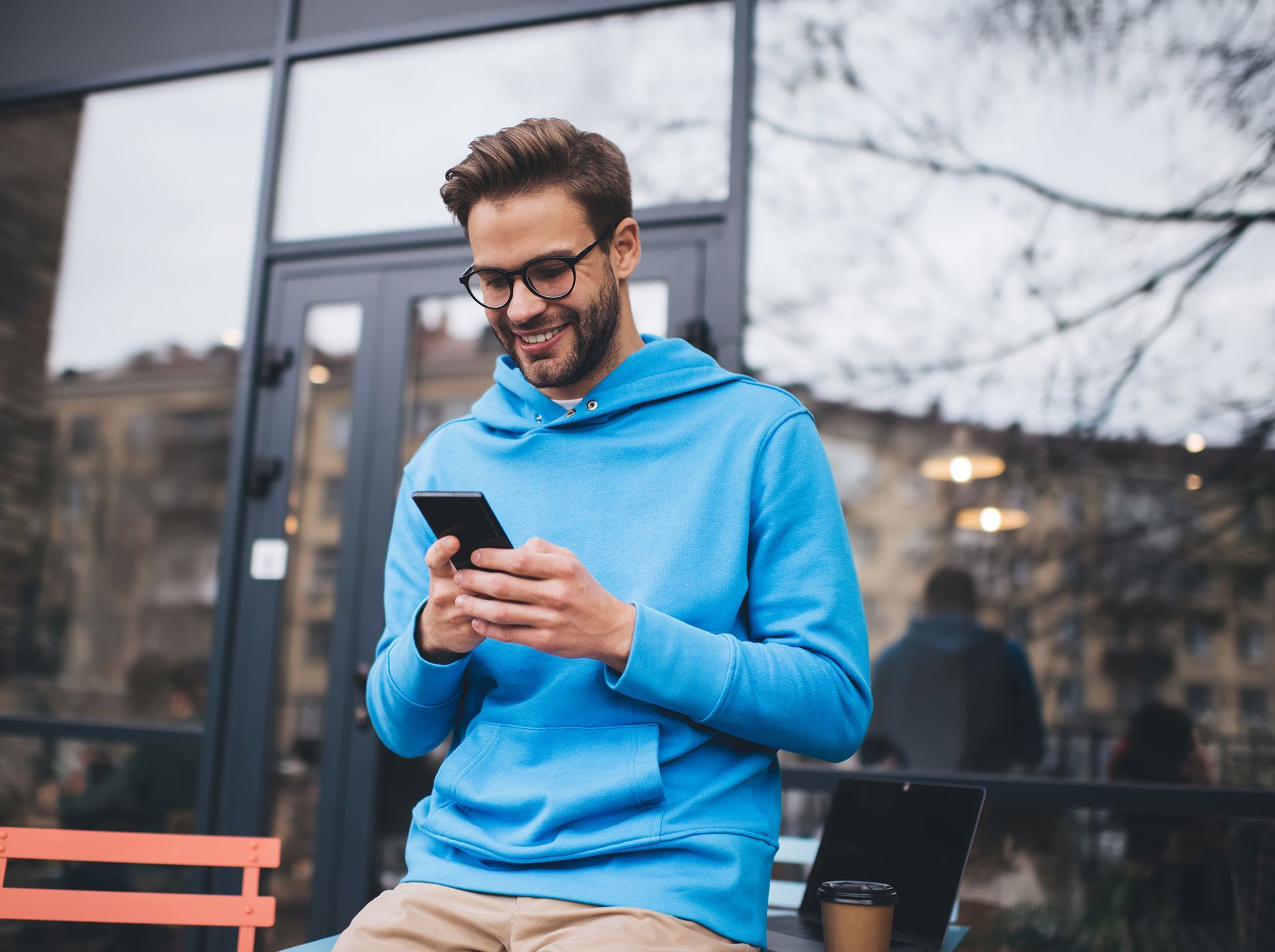 A man in a blue hoodie is sitting on a bench looking at his cell phone.