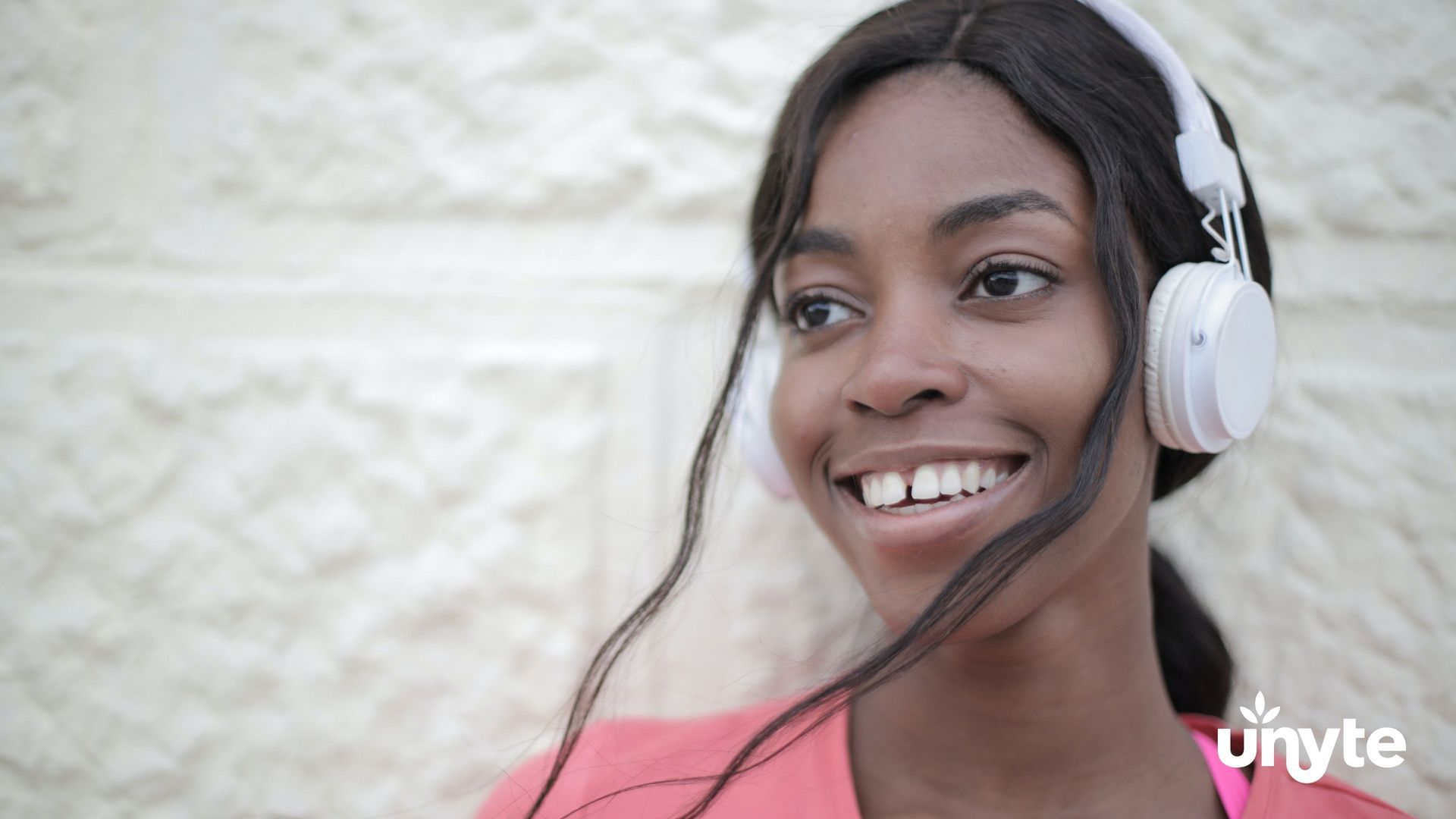 Smiling person wearing white headphones listening to Rest and Restoration Protocol outdoors against a light brick wall