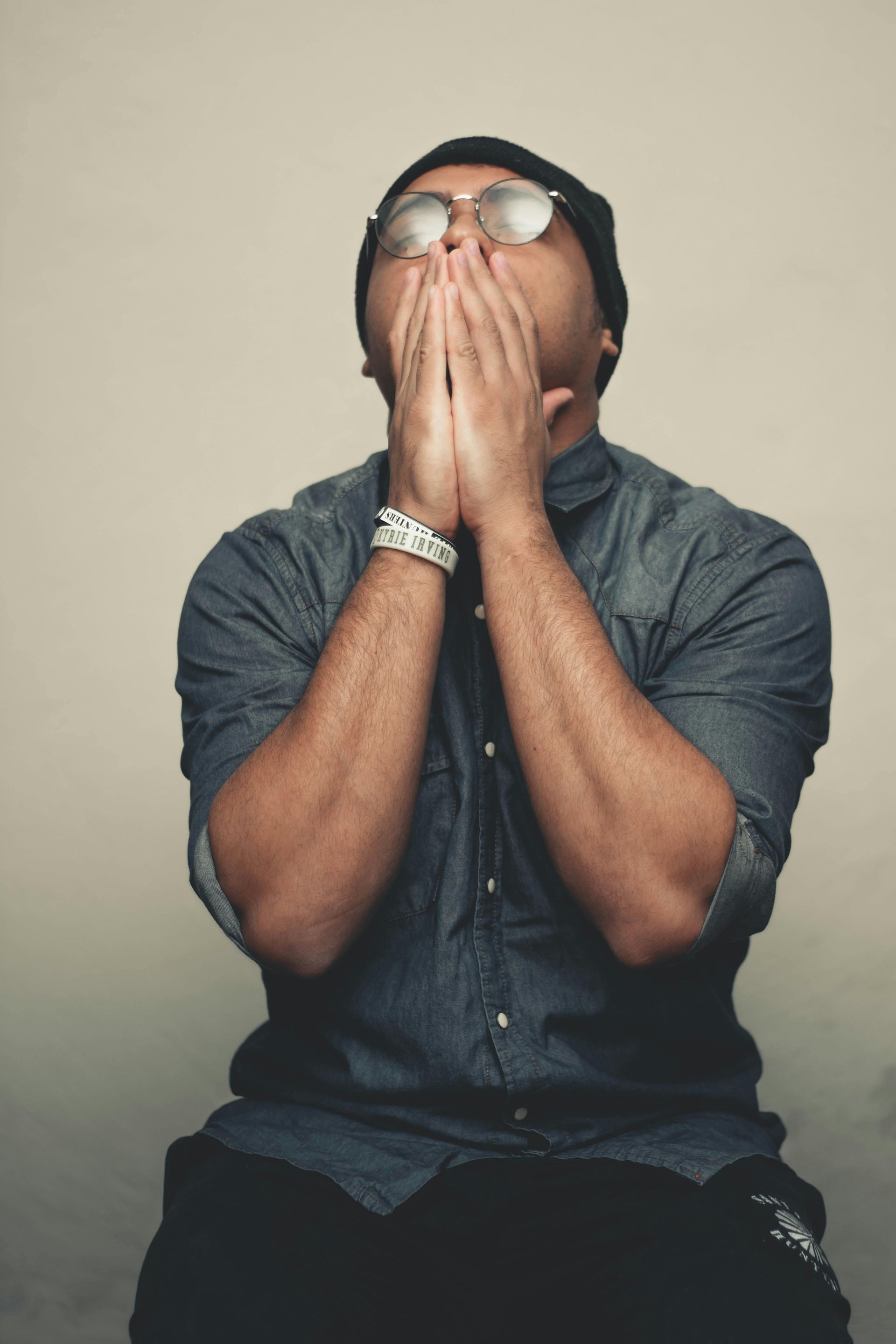 A man wearing glasses and a beanie is covering his mouth with his hands.