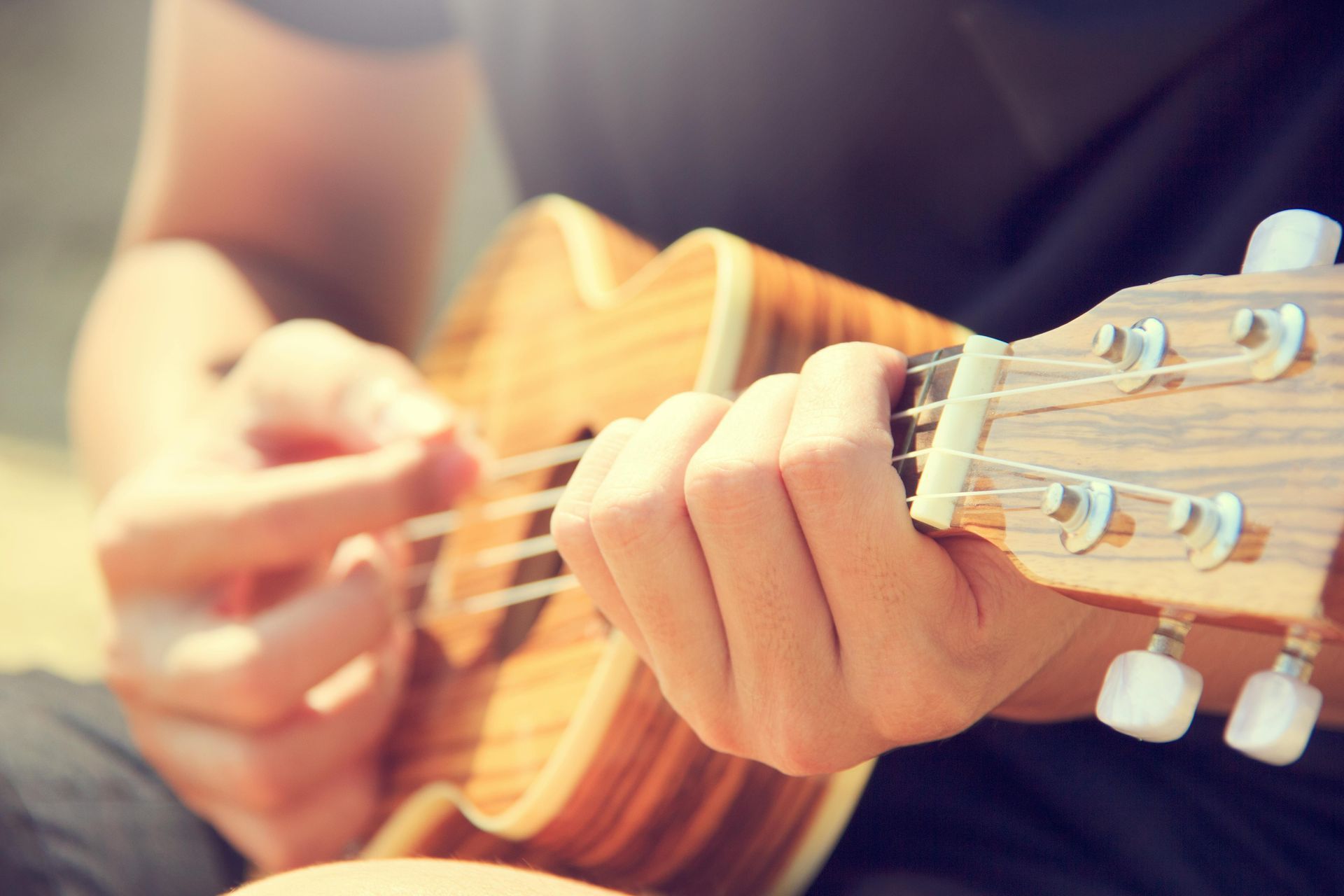 Person strumming a ukulele, close-up on hands and instrument. Brown wood, white strings, outdoor setting.