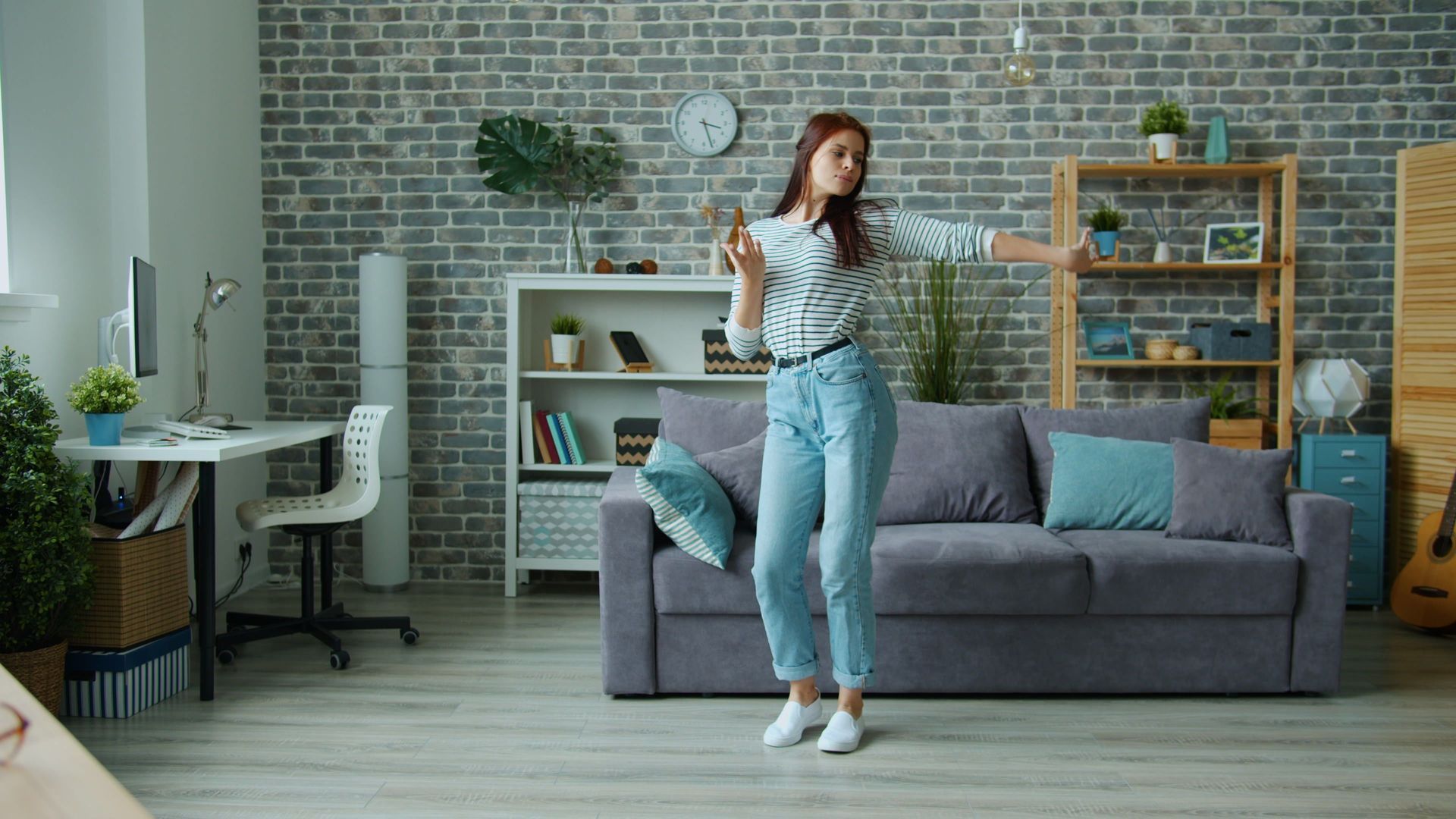 Woman dancing in a bright living room with a gray sofa and brick wall decreasing stress